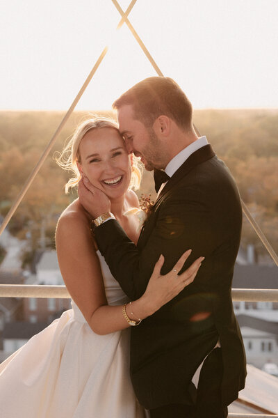 Bride smiles brightly as the groom holds her close during golden hour, sunlight glowing behind them atop a tower.
