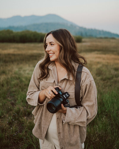 woman smiling and holding camera while standing outside with hills in the background