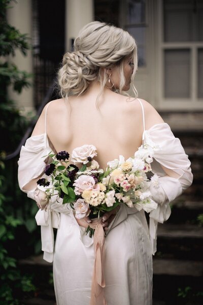 Bride in a romantic, low bun updo holding a bouquet behind her back; elegant, whimsical bridal hair styled by Klover Beauty for a Maryland wedding.