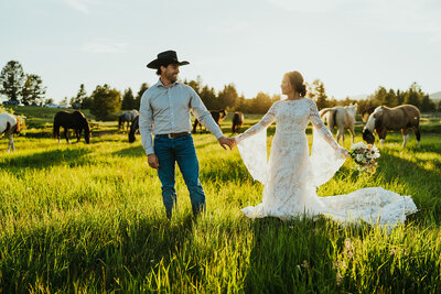 Bridal photography session where the man and woman are holding hands with their bodies facing the camera. They are looking at each other smiling, she is in her wedding dress and he is wearing a cowboy hat with a nice button up. Behind them are horses grazing.