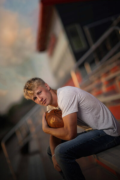 A high school senior sits on bleachers holding a football in one arm in a grey shirt and jeans at sunset