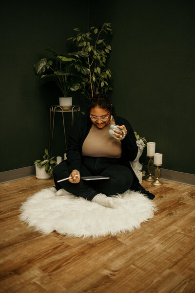Ashley sitting on the floor with an ipad in her lap, apple pencil in hand and coffee in the other.