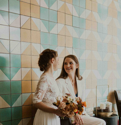 Newly wed couple holding orange flowers smile at each other in front of blue and orange tile wall