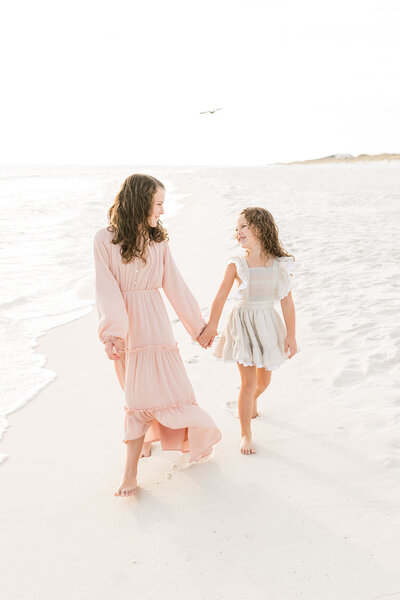 two girls walking on the beach holding their hands and smiling at each other, by Andrea Krey Photography