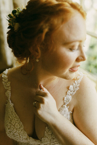 A bride with red hair in an elegant lace wedding dress looks out the window at the Red Fox Inn and Tavern in Middleburg Virginia with soft bridal makeup by kiley smith