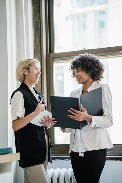 Two professional women standing and smiling during a business meeting, holding documents and engaging in a collaborative discussion in a modern office setting.