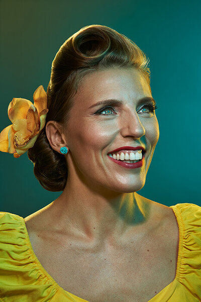 Portrait of Ylva Erevall in a vibrant yellow outfit, smiling confidently against a teal backdrop, exuding warmth and charm.