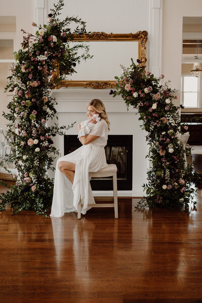 Newborn photo of mom holding baby on shoulder while sitting on a stool with a flower arch and wearing white in Annapolis Maryland.