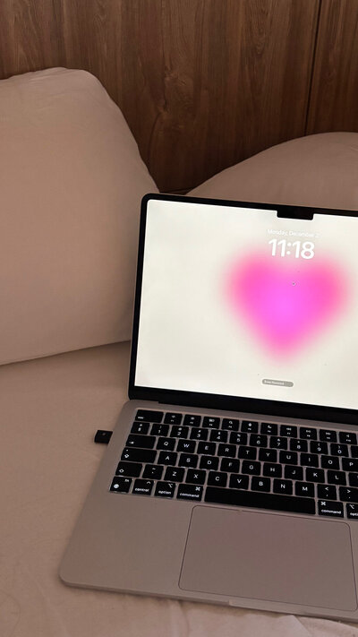 A laptop on a bed displays a lock screen with a bright pink heart and the time, 11:18. The setting feels cozy with soft lighting and wooden paneling.