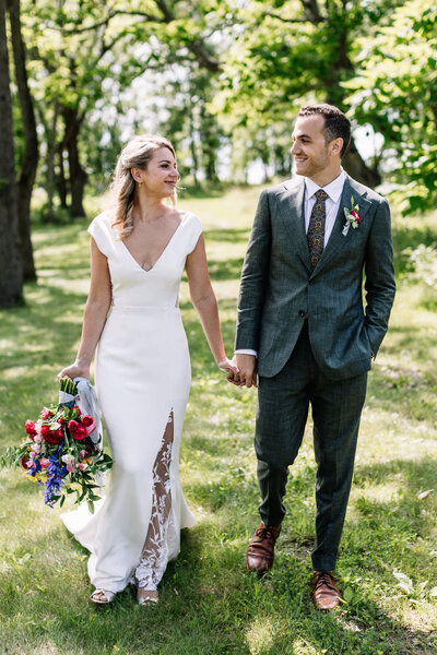 Bride and groom walk towards camera while looking at each other and smiling. Surrounded by lush green woods, bride carries colorful bouquet.