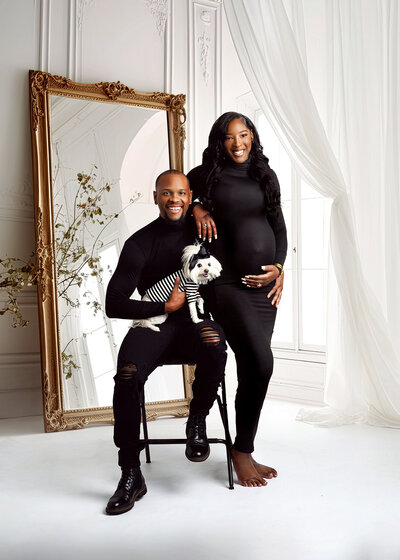 Sweet photo of expecting parents wearing black, holding their dog who is wearing black and white outfit and black bow, in a beautiful white room with large gold mirror