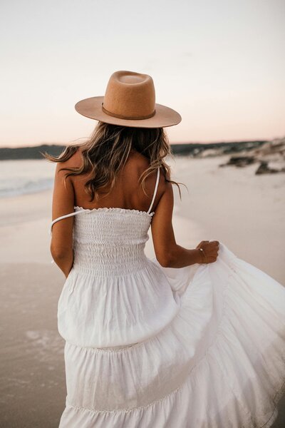 Woman in a white sundress and tan hat walking on the beach