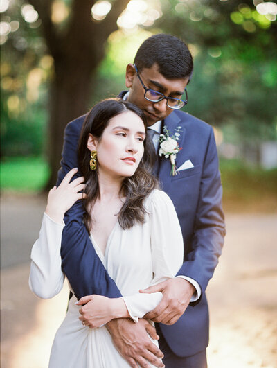 Blue eyed and black haired bride with tattoos and berry lips looks into the camera while leaning on the chest of her groom wearing a black suit in Spain.