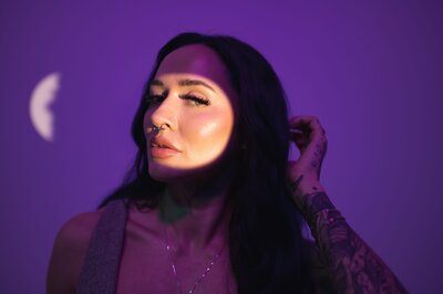 Woman with dark, long hair and tattoos on her arm side-eyes the camera with a spotlight on her face and moody purple background
