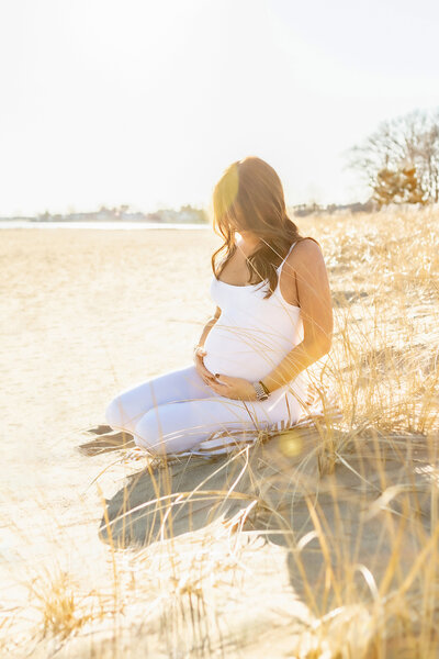 A Golden Hour maternity portrait of a mama to be wearing a white tank top and pants. Her brown hair is loosely curled and she is looking down at her belly. Sunny bokeh peeks from above her head.