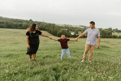 Virginia wedding photographer holding hands with her son, who is holding hands with the photographers husband as they walk through a spring wildflower field togeteher with rolling hills in the distance