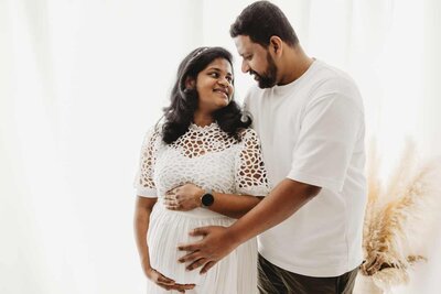 Ruua Hamdan Photography (UK): A pregnant woman in a white dress is lovingly embraced by a man.