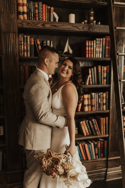 Bride and groom stand holding eachother infront of a book shelf