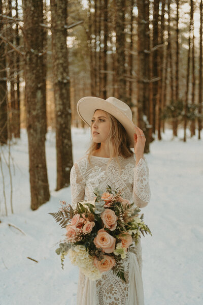 A boho bride holding the brim of her hat in a snowy forest