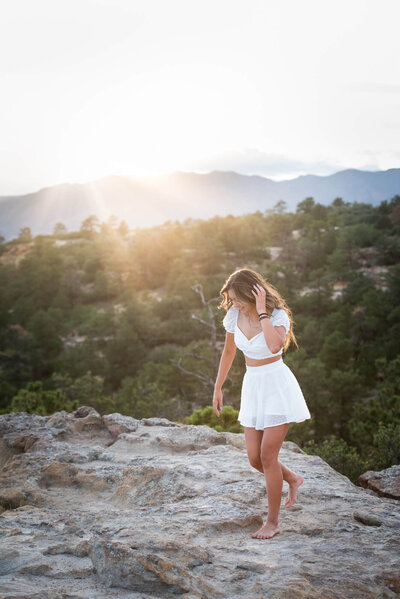 A woman in a white dress walks barefoot on a rocky overlook