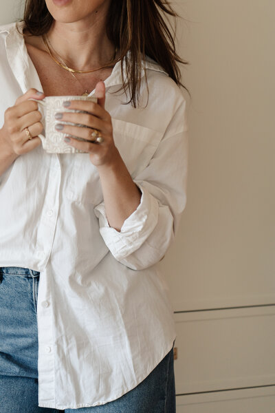Close-up of a woman in a white button-down shirt and jeans, holding a textured ceramic mug with both hands - Family Autism Support Coach at Chantal Hewitt Coaching