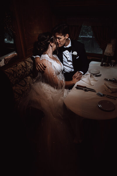 A couple sitting at a table in a dark room embraces intimately spotlighted by off camera flash