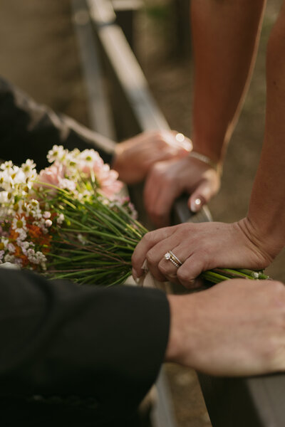 bride and groom resting hands on a fence, bride showing wedding rings and bouquet. 