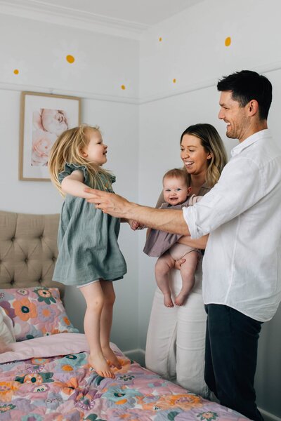 Girl jumping on bed, holding hands with family, during in-home family photoshoot