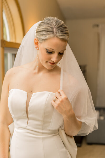 Bride in gown and veil during getting ready time on her wedding day. She's looking down as she holds her veil with her wedding ring on. 