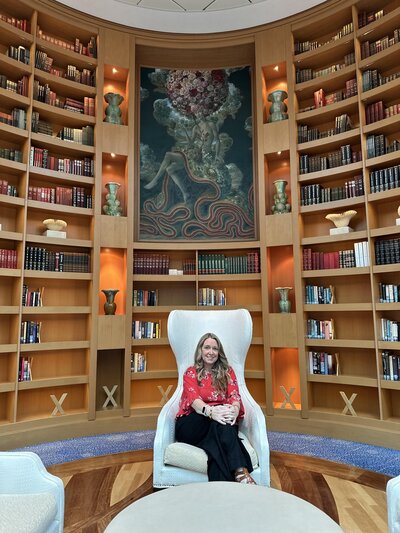 Terra from Adventures by Terra sitting in a white chair wearing a red top and black pants with the walls of library books behind on the Celebrity Equinox cruise ship