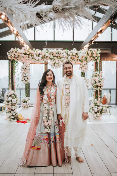 A couple smiles under a floral canopy, dressed in elegant traditional attire during their wedding ceremony at Jersey City Hyatt House