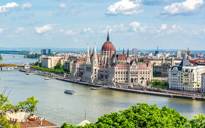 An aerial view of Budapest's Parliament building along the Danube River showcases its iconic gothic architecture, complete with spires and a central dome. Boats drift by as the cityscape stretches into the distance under a partly cloudy sky, capturing the essence of epic travel moments.