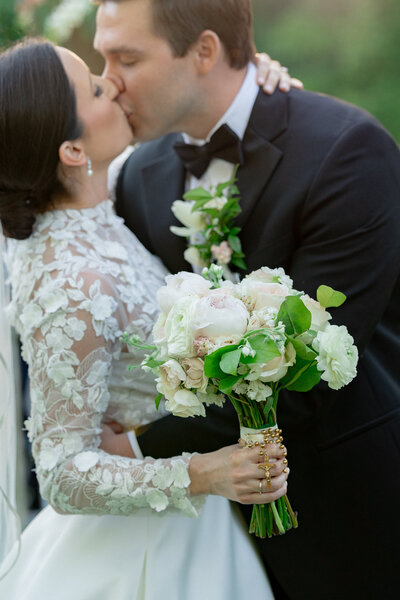 woman in lace white dress and man in tux kissing with a bouquet of flowers