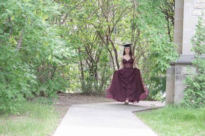 Girl in a burgundy dress walking forward wearing a grad cap at the university grounds in Saskatoon.