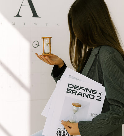 woman holding an hourglass besides a calendar whilst holding a branding workbook in her hands