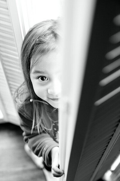 A little girl looks straight at the camera with a shy smile and soulful eyes and with her face half-hidden by a door 