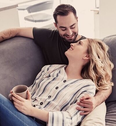 A couple smile at one another as they rest on a sofa. The man holds an arm around his partner as she holds a coffee mug. This could represent a couple reconnecting after healing from infidelity. An affair recovery therapist in Florida can help with your infidelity recovery. We offer affair counseling in Florida and other services including an affair recovery program.