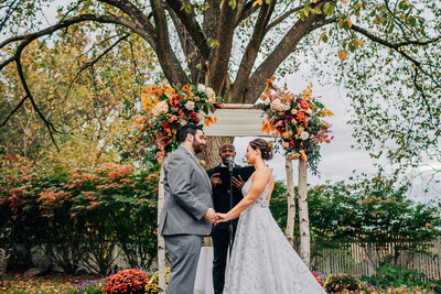 A bride, groom and officiant stand under a chuppah with autumn flowers