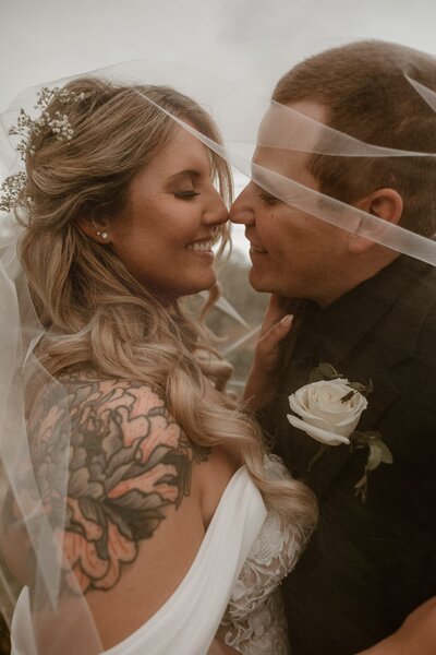 Close-up of a bride and groom intimately sharing a veil-covered moment, with the groom's boutonniere and the bride's tattoo visible, at Locust Grove Brewery.
