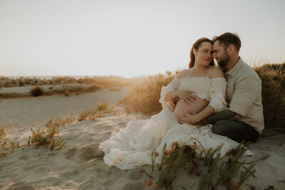 Image of a family of three, holding hands and walkin along a beach.  The mother is pregnant and wearing a flowing white gown. 