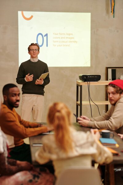 Corporate training session with presenter speaking to engaged audience, projector displaying content, representing professional development workshop.