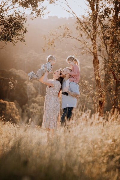 Family in a field at golden hour , daughter is on dads shoulders, son is being lifted into the air by mum.