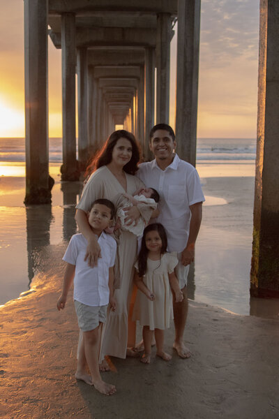 Family of four standing beneath a pier at sunset, with the parents holding their children