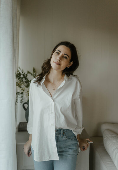 Allison Stoddard wearing a white button down and blue jeans looking at camera while leaning back on dresser with books and plants