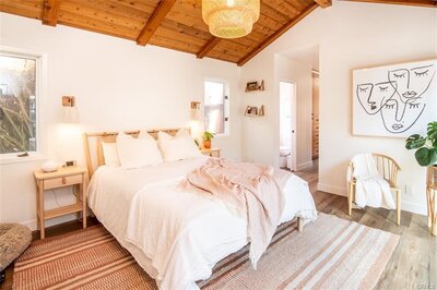 Boho bedroom with vaulted wooden ceiling, bright white walls, light wood furniture, crisp white bedding, cozy chairs, and modern wall art.