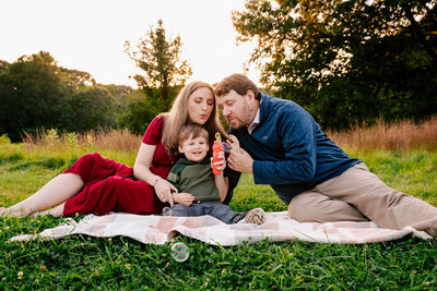 Young mother and father sit on a blanket in a meadow, blowing bubbles with their toddler. He sits between them and is giggling.