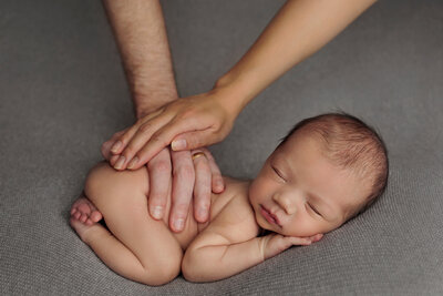 A newborn baby is peacefully sleeping on a soft gray blanket, lying on their side with their tiny hands tucked under their chin. Two adult hands, one with a wedding ring, gently rest on the baby's back, providing support and a sense of security. The baby's expression is calm and content, and the scene exudes warmth and tenderness. The soft lighting and neutral background keep the focus on the baby's serene face and the comforting presence of the adult hands, making this a heartwarming and intimate portrait.