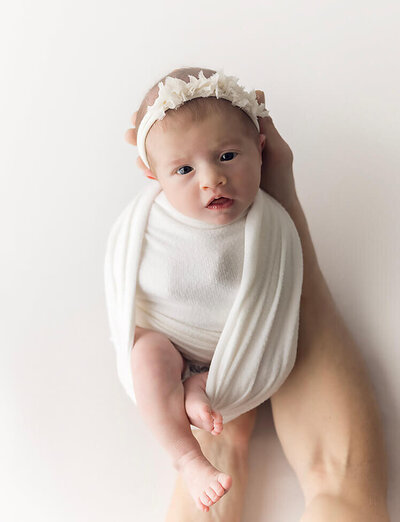 A wrapped newborn baby being held by daddy's hand looks into the camera.