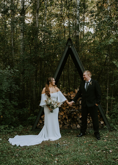 Bride and groom standing in front of a wood pile in the woods.