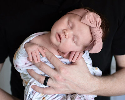 Newborn girl wearing a pink headband sleeping in her father's hands during her lifestyle newborn photo shoot.